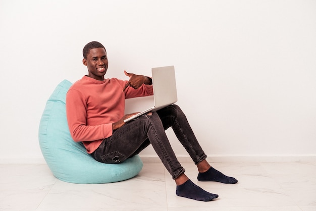 Jeune homme afro-américain assis sur une bouffée à l'aide d'un ordinateur portable isolé sur fond blanc personne pointant à la main vers un espace de copie de chemise, fier et confiant