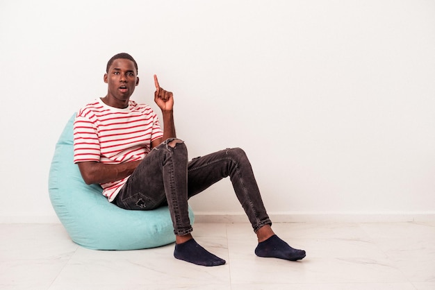 Photo jeune homme afro-américain assis sur une bouffée isolé sur fond blanc ayant une bonne idée, concept de créativité.