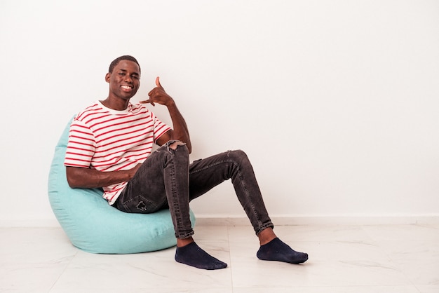 Jeune Homme Afro-américain Assis Sur Une Bouffée Isolée Sur Fond Blanc Montrant Un Geste D'appel De Téléphone Portable Avec Les Doigts.