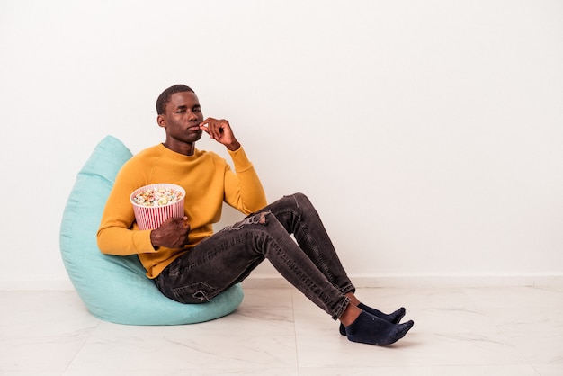 Jeune homme afro-américain assis sur une bouffée mangeant du pop-corn isolé sur fond blanc avec les doigts sur les lèvres gardant un secret.