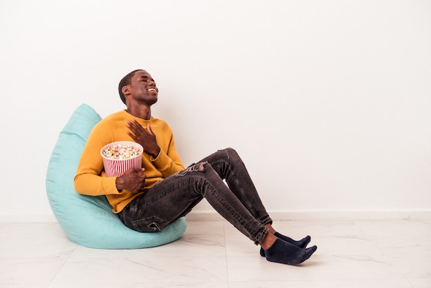 Jeune homme afro-américain assis sur une bouffée mangeant du pop-corn isolé sur fond blanc rit fort en gardant la main sur la poitrine.