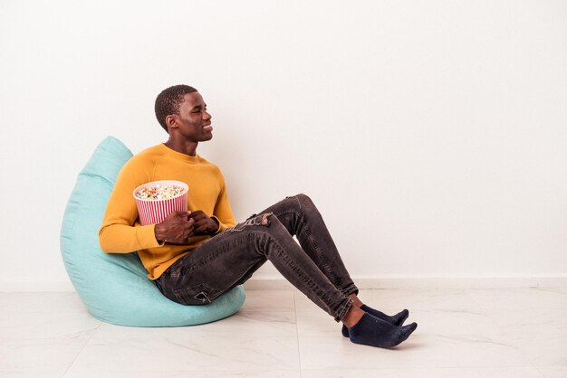 Jeune homme afro-américain assis sur une bouffée manger du pop-corn isolé sur fond blanc regarde de côté souriant joyeux et agréable