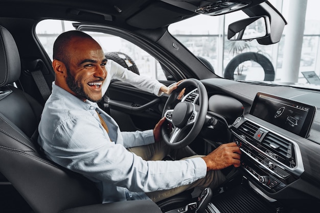 Jeune homme afro-américain assis dans une nouvelle voiture dans une salle d'exposition de voitures et regardant autour