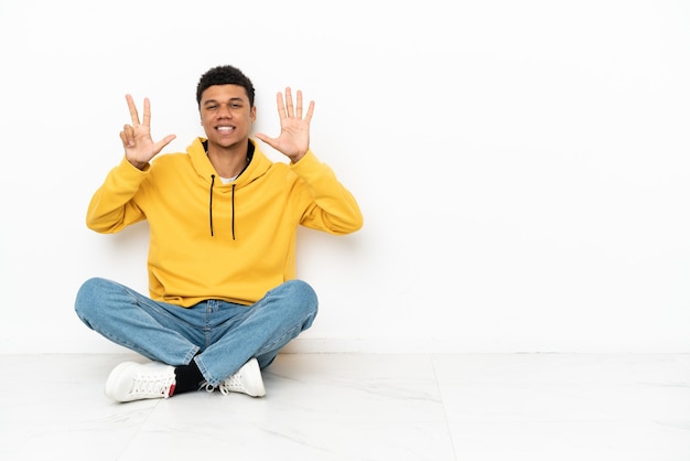 Jeune homme afro-américain assis sur le sol isolé sur fond blanc comptant huit avec les doigts