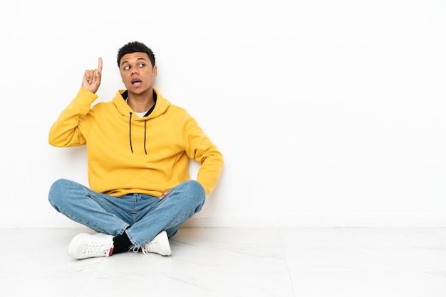 Jeune homme afro-américain assis sur le sol isolé sur fond blanc dans l'intention de réaliser la solution tout en levant un doigt vers le haut