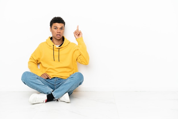 Jeune homme afro-américain assis sur le sol isolé sur fond blanc pensant à une idée pointant le doigt vers le haut