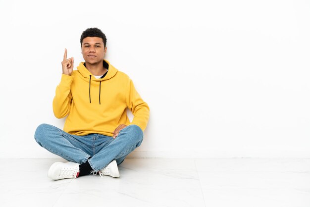 Jeune homme afro-américain assis sur le sol isolé sur fond blanc pointant avec l'index une excellente idée