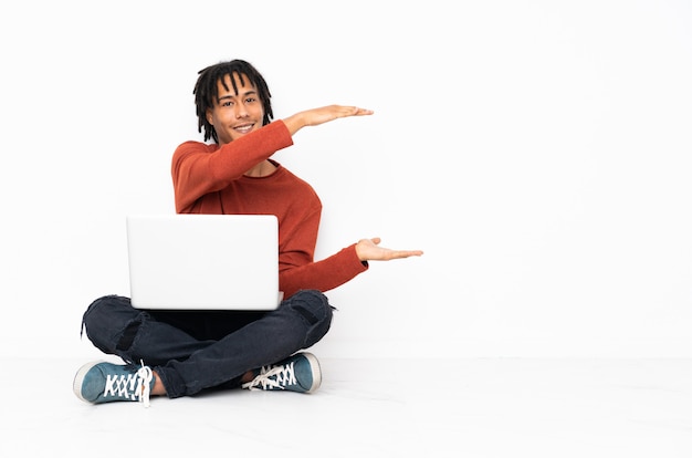 Jeune homme afro-américain assis sur le sol et travaillant avec son ordinateur portable tenant copyspace pour insérer une annonce