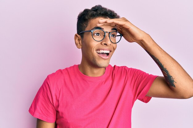 Jeune homme afro-américain beau portant des lunettes sur fond rose très heureux et souriant regardant loin avec la main sur la tête recherche concept