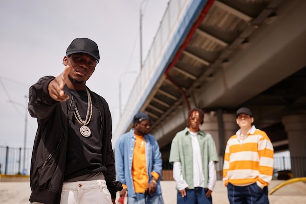 Jeune homme afro-américain en casquette noire et veste de bombardier vous pointant du doigt alors qu'il se tient contre un groupe d'amis interculturels à l'extérieur