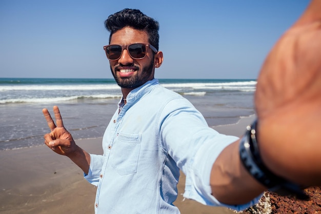 Jeune homme afro-américain en chemise bleue et jeans faisant un portrait de selfie sur la caméra frontale d'un smartphone sur la plage de l'océan