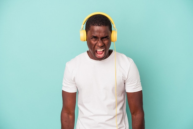 Jeune homme afro-américain écoutant de la musique isolé sur fond bleu criant très en colère et agressif.