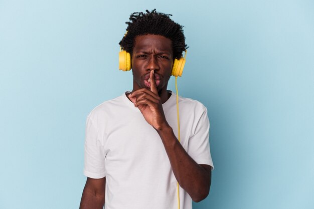 Jeune homme afro-américain écoutant de la musique isolée sur fond bleu gardant un secret ou demandant le silence.