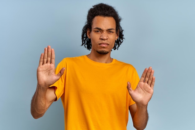 Jeune homme afro-américain avec une expression effrayée sur son visage faisant un geste effrayé avec les paumes comme s'il essayait de se défendre de quelqu'un, demandant d'arrêter, gesticulant avec les mains sur fond bleu