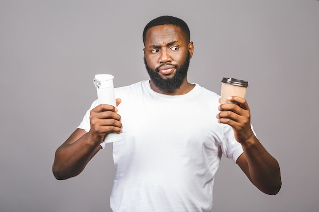 Jeune homme afro-américain faisant choisir entre une tasse de café et recycler un debout sur fond gris isolé.