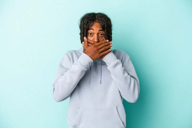 Jeune homme afro-américain isolé sur fond bleu couvrant la bouche avec les mains à l'air inquiet.