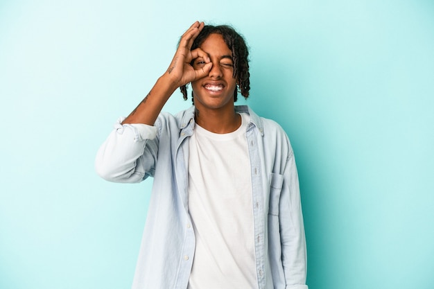 Jeune homme afro-américain isolé sur fond bleu excité en gardant le geste ok sur les yeux.