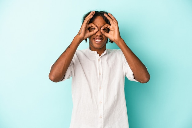 Jeune homme afro-américain isolé sur fond bleu excité en gardant le geste ok sur les yeux.