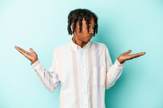 Jeune homme afro-américain isolé sur fond bleu, haussant les épaules confus et douteux pour tenir un espace de copie.