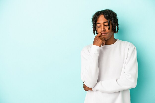 Jeune homme afro-américain isolé sur fond bleu regardant de côté avec une expression douteuse et sceptique.