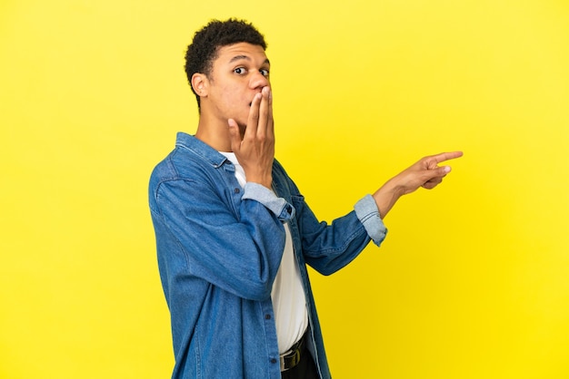 Jeune homme afro-américain isolé sur fond jaune avec une expression de surprise tout en pointant le côté