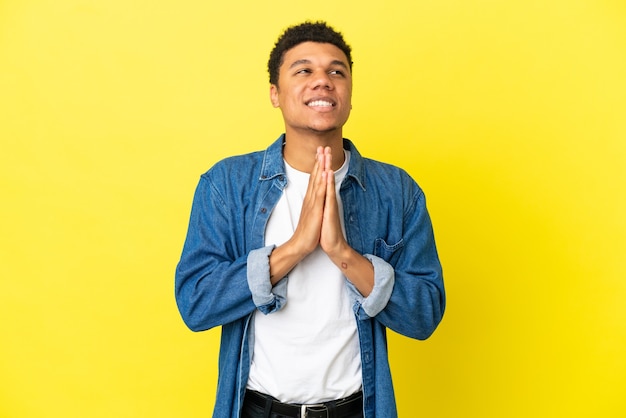 Jeune homme afro-américain isolé sur fond jaune maintient la paume ensemble. La personne demande quelque chose