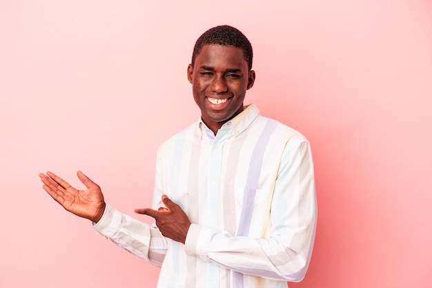 Jeune homme afro-américain isolé sur fond rose excité tenant un espace de copie sur la paume