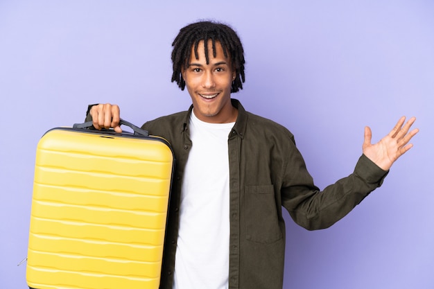 Jeune homme afro-américain isolé sur violet en vacances avec valise de voyage et surpris