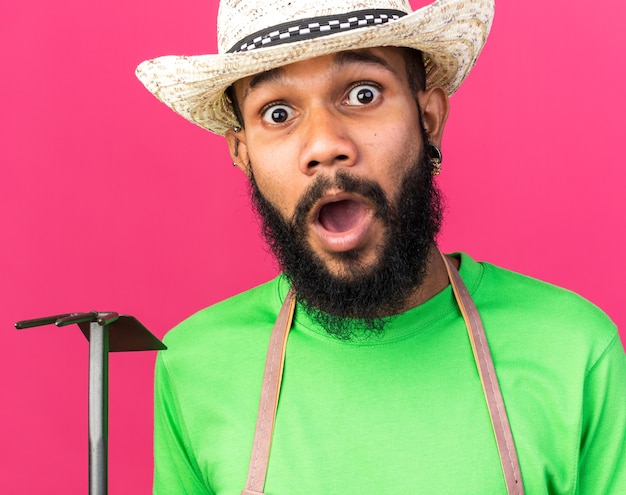 Jeune homme afro-américain de jardinier surpris portant un chapeau de jardinage tenant un râteau à houe isolé sur un mur rose