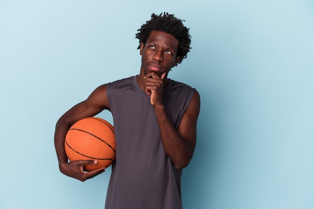 Jeune Homme Afro-américain Jouant Au Basket-ball Isolé Sur Fond Bleu Regardant De Côté Avec Une Expression Douteuse Et Sceptique.