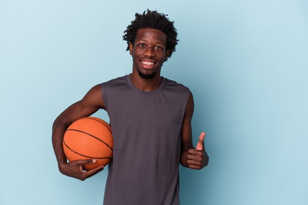 Jeune homme afro-américain jouant au basket-ball isolé sur fond bleu souriant et levant le pouce vers le haut