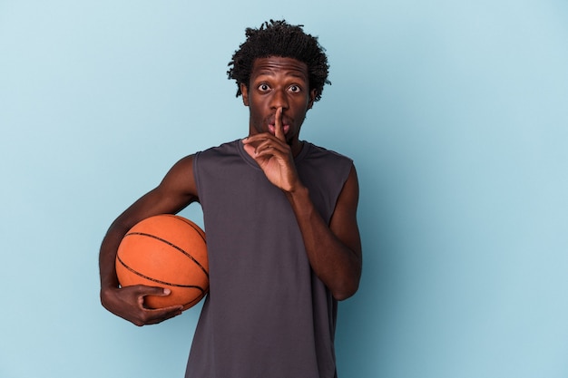 Jeune homme afro-américain jouant au basket isolé sur fond bleu gardant un secret ou demandant le silence.