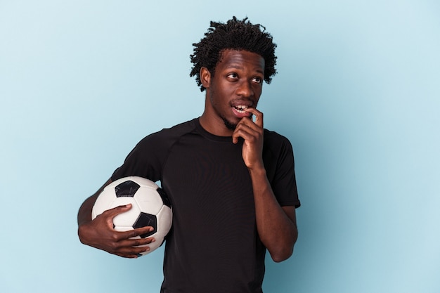 Jeune homme afro-américain jouant au football isolé sur fond bleu détendu en pensant à quelque chose en regardant un espace de copie.