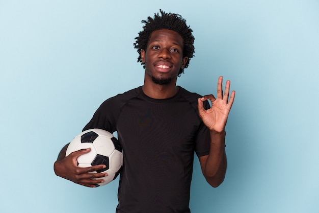 Jeune homme afro-américain jouant au football isolé sur fond bleu joyeux et confiant montrant un geste correct.