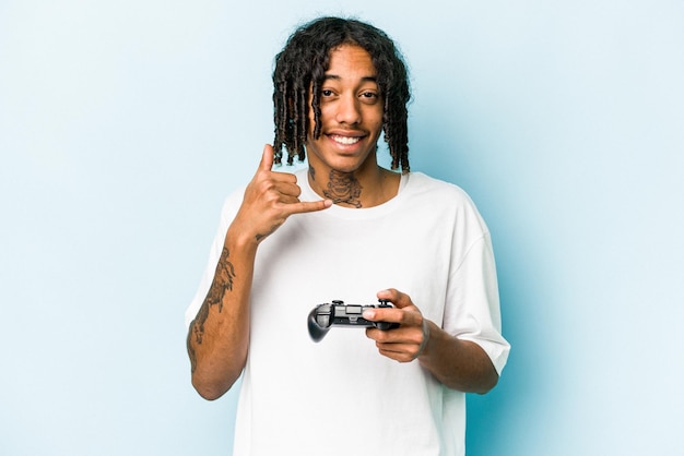 Jeune homme afro-américain jouant avec un contrôleur de jeu vidéo isolé sur fond bleu montrant un geste d'appel de téléphone portable avec les doigts