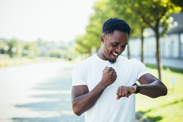 Un jeune homme afro-américain, lors d'un jogging matinal dans un T-shirt blanc dans le parc, satisfait du résultat d'un entraînement engagé dans un mode de vie actif de remise en forme