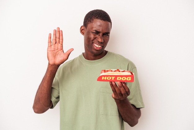 Jeune homme afro-américain mangeant un hot-dog isolé sur fond blanc souriant joyeux montrant le numéro cinq avec les doigts