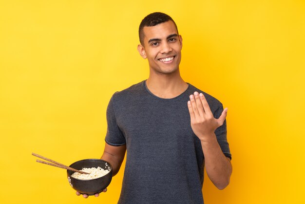 Jeune homme afro-américain sur mur jaune isolé invitant à venir avec la main. Heureux que tu sois venu en tenant un bol de nouilles avec des baguettes