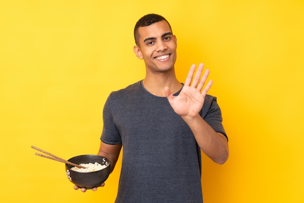 Jeune homme afro-américain sur mur jaune isolé saluant avec la main avec une expression heureuse tout en tenant un bol de nouilles avec des baguettes