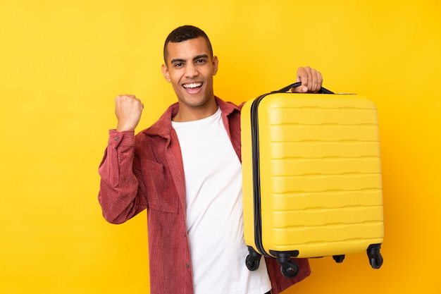 Jeune homme afro-américain sur mur jaune isolé en vacances avec valise de voyage et un chapeau