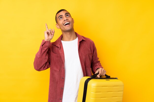 Jeune homme afro-américain sur mur jaune isolé en vacances avec valise de voyage et pointant vers le haut