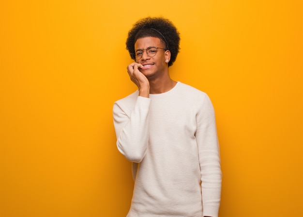 Jeune homme afro-américain sur un mur orange se ronger les ongles, nerveux et très anxieux