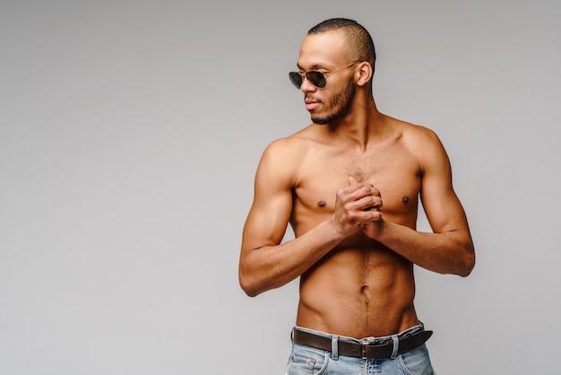 Jeune homme afro-américain musclé torse nu portant des lunettes de soleil sur un mur gris clair