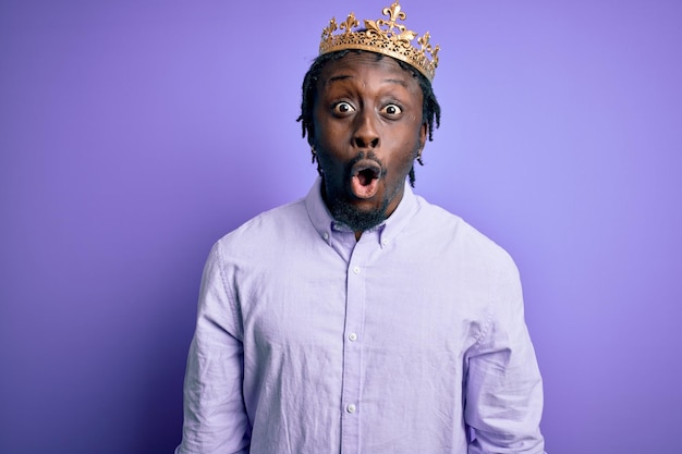 Jeune homme afro-américain portant la couronne dorée du roi sur fond violet isolé effrayé et choqué par la peur de l'expression surprise et le visage excité