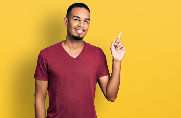 Jeune homme afro-américain portant un t-shirt décontracté souriant avec une idée ou une question pointant le doigt vers le haut avec un visage heureux numéro un