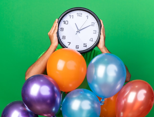 Jeune homme afro-américain portant un t-shirt vert debout derrière des ballons recouverts d'une horloge murale isolée sur un mur vert