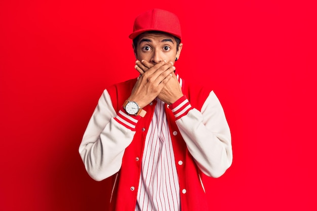 Jeune homme afro-américain portant un uniforme de baseball choqué couvrant la bouche avec les mains pour un concept secret d'erreur