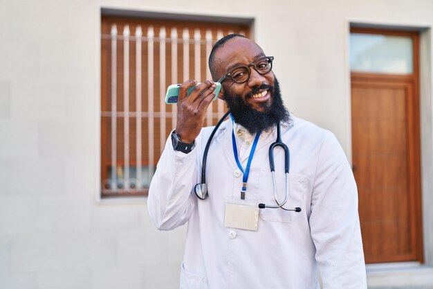 Jeune homme afro-américain portant un uniforme de médecin écoutant un message audio par le smartphone à l'hôpital