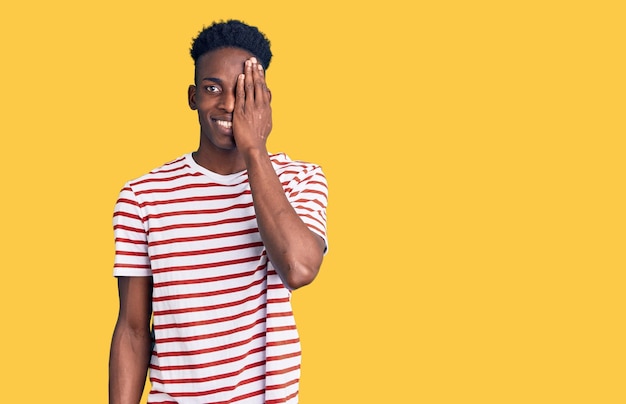 Jeune homme afro-américain portant des vêtements décontractés couvrant un œil avec la main, un sourire confiant sur le visage et une émotion surprise.