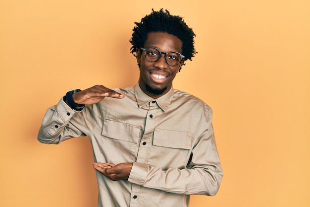 Photo jeune homme afro-américain portant des vêtements décontractés et des lunettes gesticulant avec les mains montrant le symbole de mesure de signe de grande et grande taille souriant en regardant le concept de mesure de la caméra